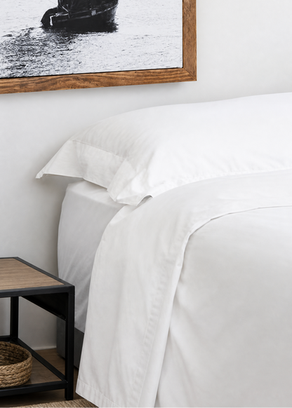 A close-up of a neatly made bed with crisp white bedding, positioned next to a small dark wood side table. On the wall above the bed hangs a large framed black-and-white photograph of a sailboat.