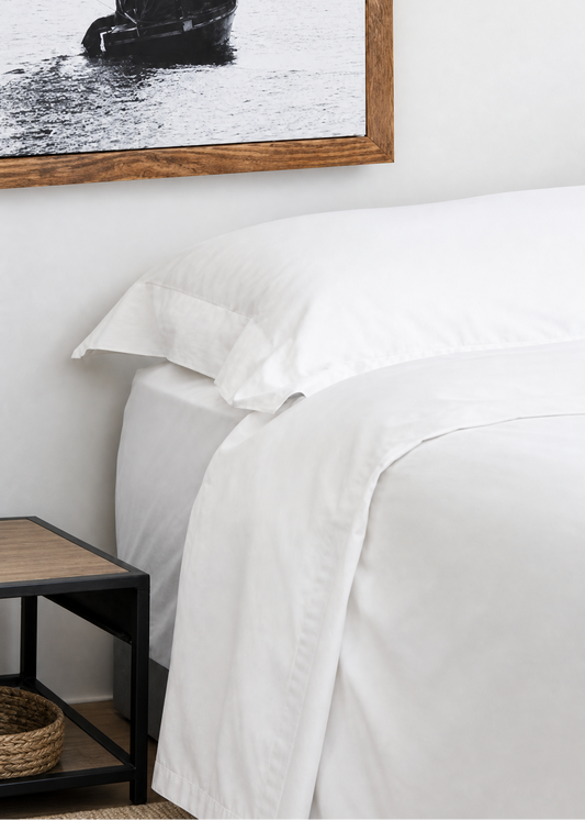 A close-up of a neatly made bed with crisp white bedding, positioned next to a small dark wood side table. On the wall above the bed hangs a large framed black-and-white photograph of a sailboat.