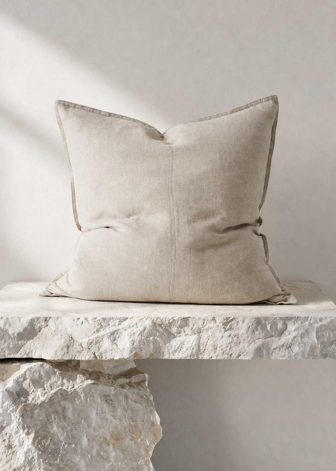 A square beige linen throw pillow with a subtle stitched center seam, resting on a textured white stone ledge against a neutral gray wall.