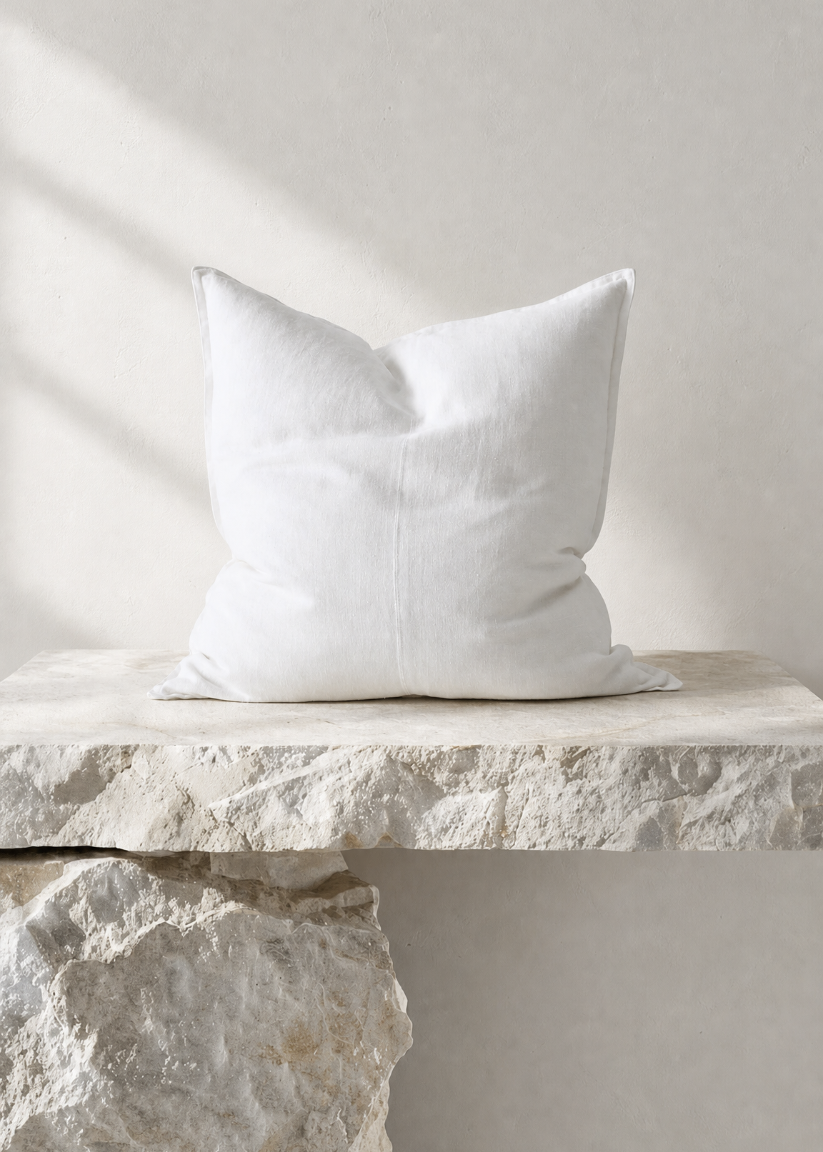 A white square throw pillow with a subtle stitched center seam, resting on a rustic stone ledge against a textured light gray wall with soft diagonal shadows.
