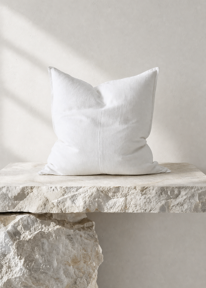 A white square throw pillow with a subtle stitched center seam, resting on a rustic stone ledge against a textured light gray wall with soft diagonal shadows.