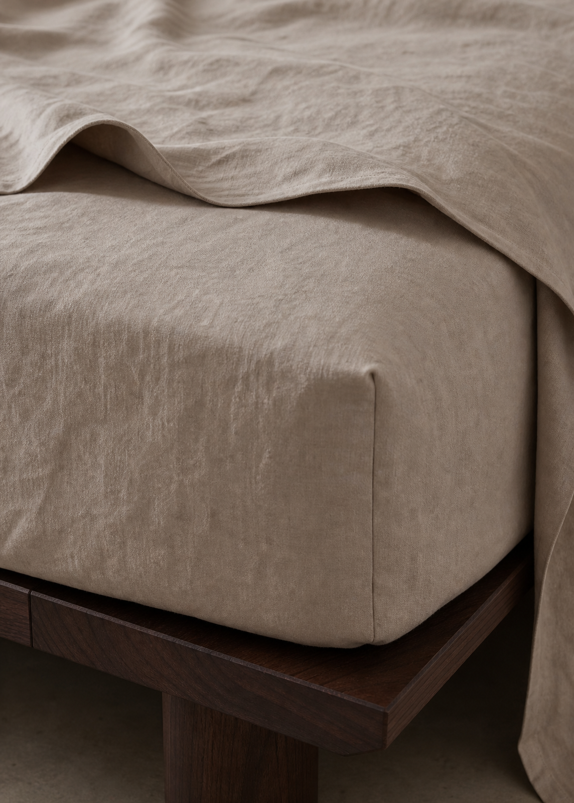 A close-up of a beige linen bed sheet or duvet draped over a bed frame with a dark wood base, set against a neutral concrete floor.