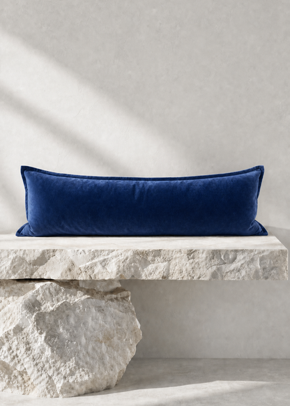 A long rectangular navy blue velvet pillow displayed on a rustic white stone bench against a textured light gray wall.