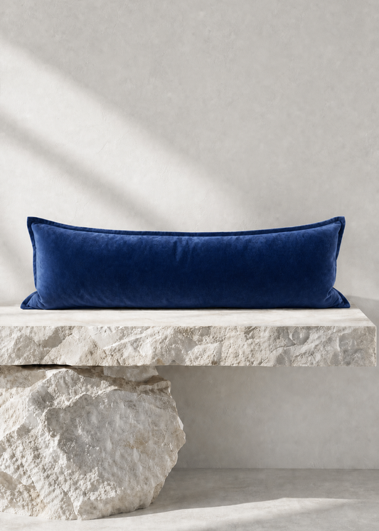 A long rectangular navy blue velvet pillow displayed on a rustic white stone bench against a textured light gray wall.