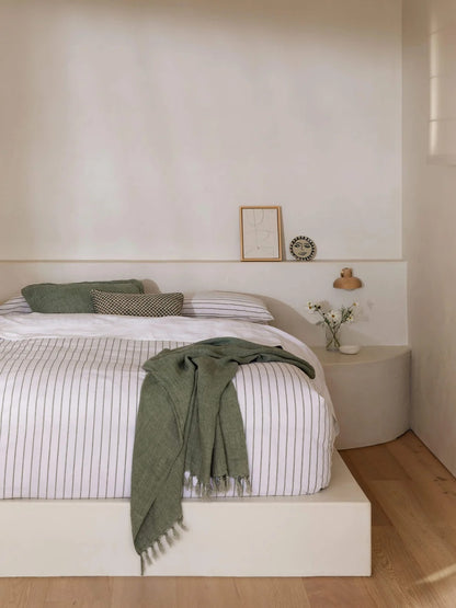 A minimalist bedroom scene featuring a platform bed with white bedding and thin vertical gray stripes, styled with olive green pillows and a matching olive green throw blanket. The room has warm off-white walls, light wood flooring, and a small side table with a lamp and vase.