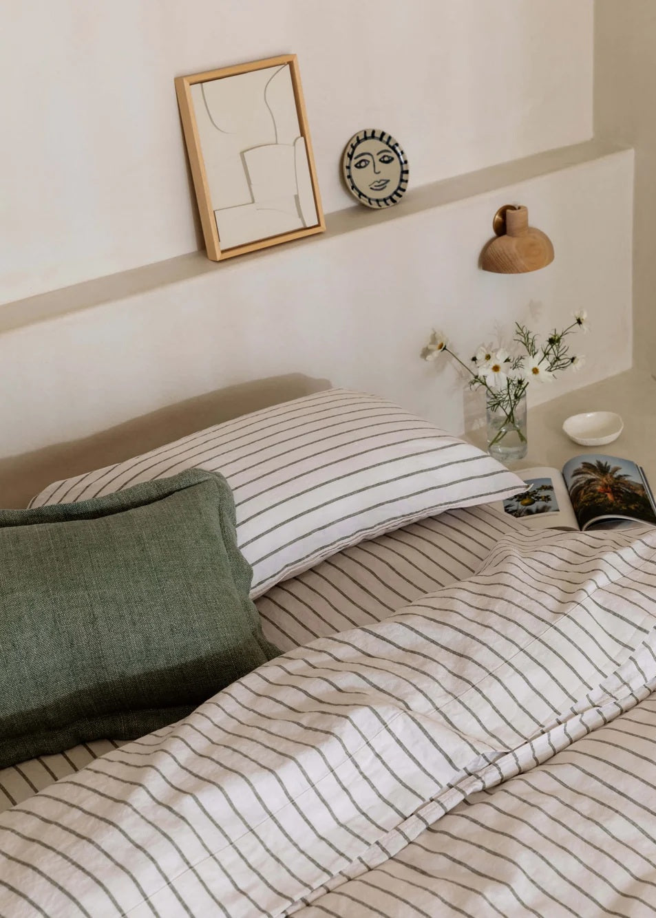 A styled bedroom scene showing a bed with cream bedding featuring thin dark vertical stripes, a dark green textured pillow, and a matching headboard. Decorative wall art, a small wall clock, a wooden lamp, and a vase of white flowers are arranged on the wall above the bed.