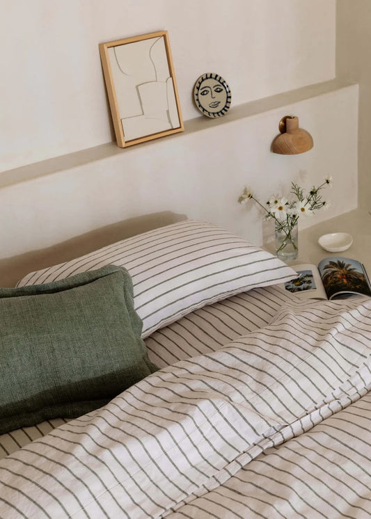 A styled bedroom scene showing a bed with cream bedding featuring thin dark vertical stripes, a dark green textured pillow, and a matching headboard. Decorative wall art, a small wall clock, a wooden lamp, and a vase of white flowers are arranged on the wall above the bed.