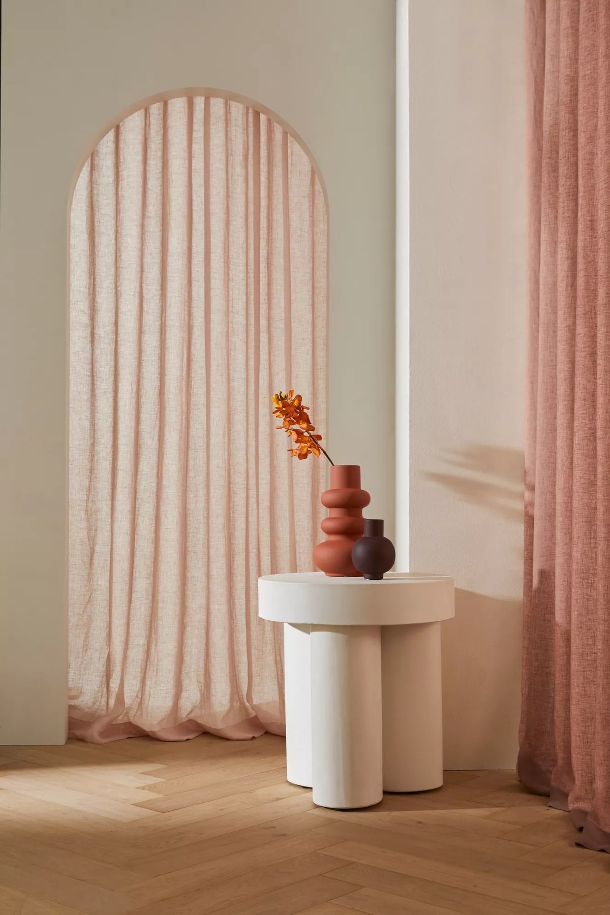 A styled interior scene with a white sculptural side table holding a terracotta vase and a small dark vase, set against a warm beige wall with an arched window draped in blush pink fabric and a matching curtain on the right.