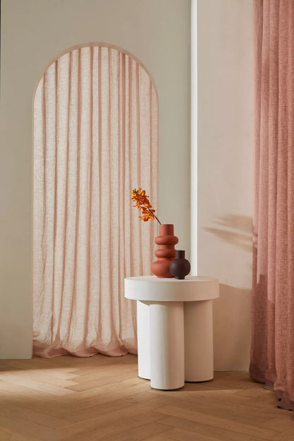 A styled interior scene with a white sculptural side table holding a terracotta vase and a small dark vase, set against a warm beige wall with an arched window draped in blush pink fabric and a matching curtain on the right.