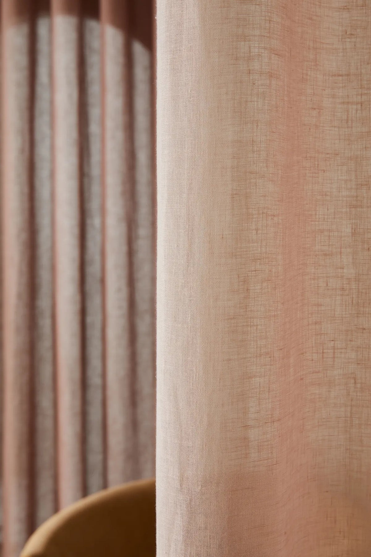 A close-up of a textured linen curtain in a dusty peach-pink color, with a matching pleated curtain panel blurred in the background.