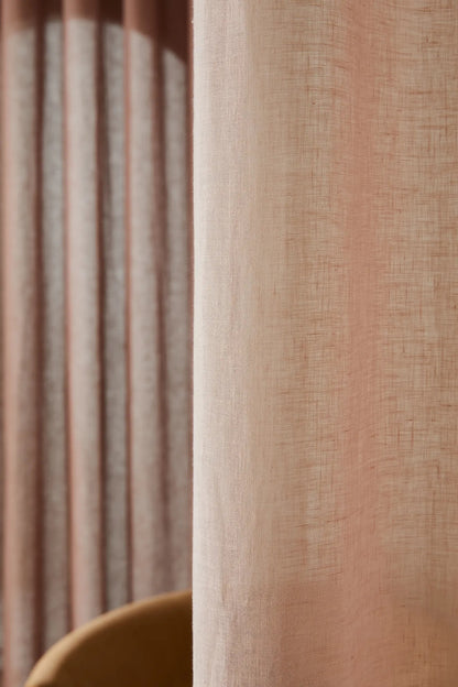 A close-up of a textured linen curtain in a dusty peach-pink color, with a matching pleated curtain panel blurred in the background.