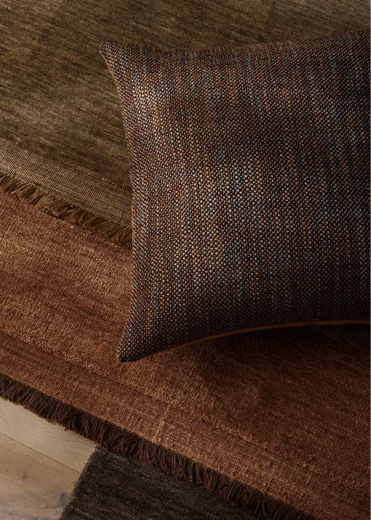 A close-up of a textured throw pillow in a dark brown and bronze woven fabric, resting on a rust-brown fringed rug over a neutral surface.