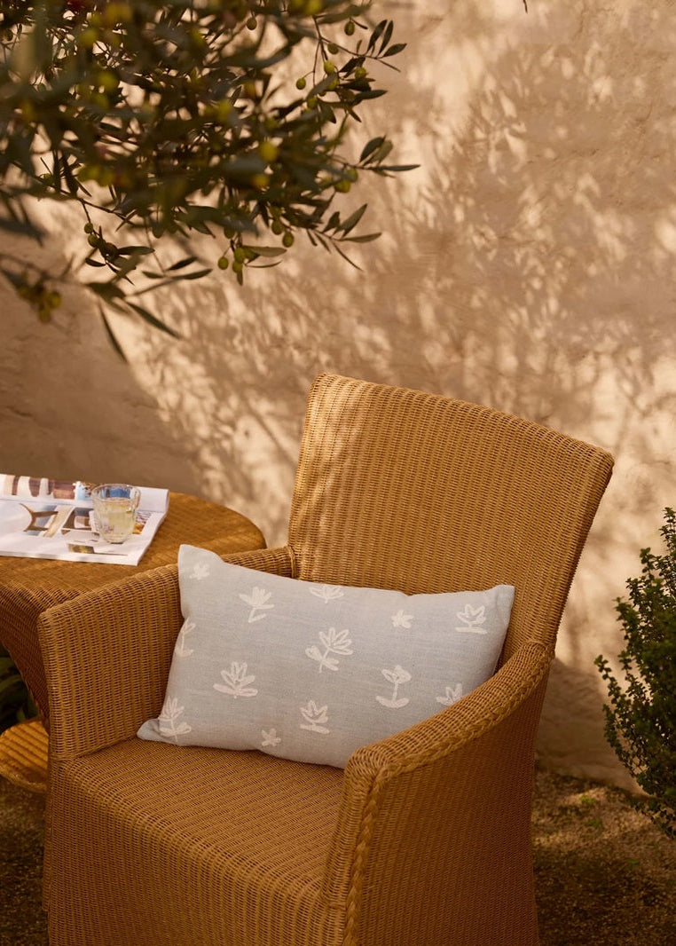 A light blue rectangular throw pillow with a white botanical pattern rests on a tan wicker armchair in an outdoor patio setting with warm sunlight and shadows.