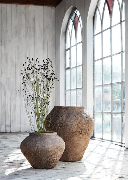 Two large stone vases with dried plants on a wooden floor in a room with large windows.