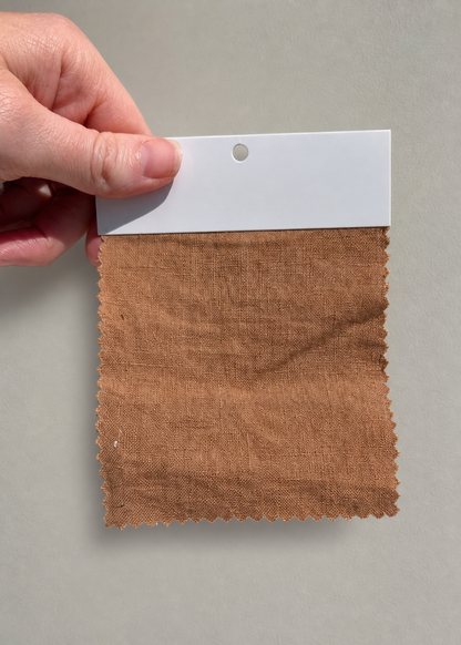 A hand holding a fabric swatch card showing a textured brown linen-like material against a plain light gray background.