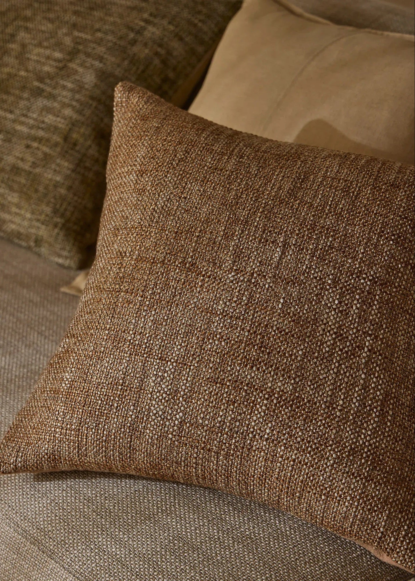 A square brown woven throw pillow with a textured linen-like fabric, resting on a beige upholstered sofa next to other pillows.