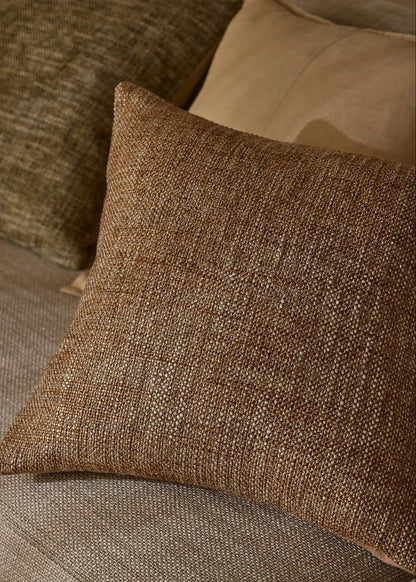 A square brown woven throw pillow with a textured linen-like fabric, resting on a beige upholstered sofa next to other pillows.