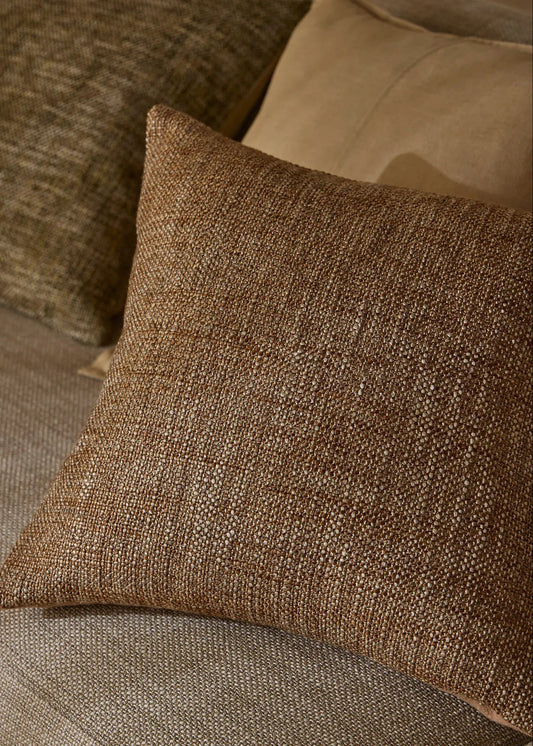 A square brown woven throw pillow with a textured linen-like fabric, resting on a beige upholstered sofa next to other pillows.