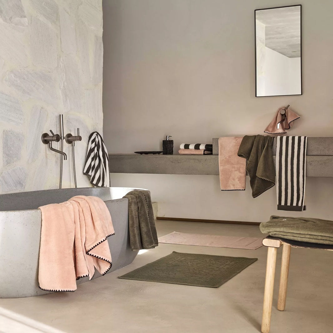 A modern bathroom scene with a gray freestanding bathtub draped in soft pink and dark gray towels, a floating shelf with folded linens and a mirror, and neutral-toned textiles throughout.