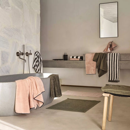 A modern bathroom scene with a gray freestanding bathtub draped in soft pink and dark gray towels, a floating shelf with folded linens and a mirror, and neutral-toned textiles throughout.