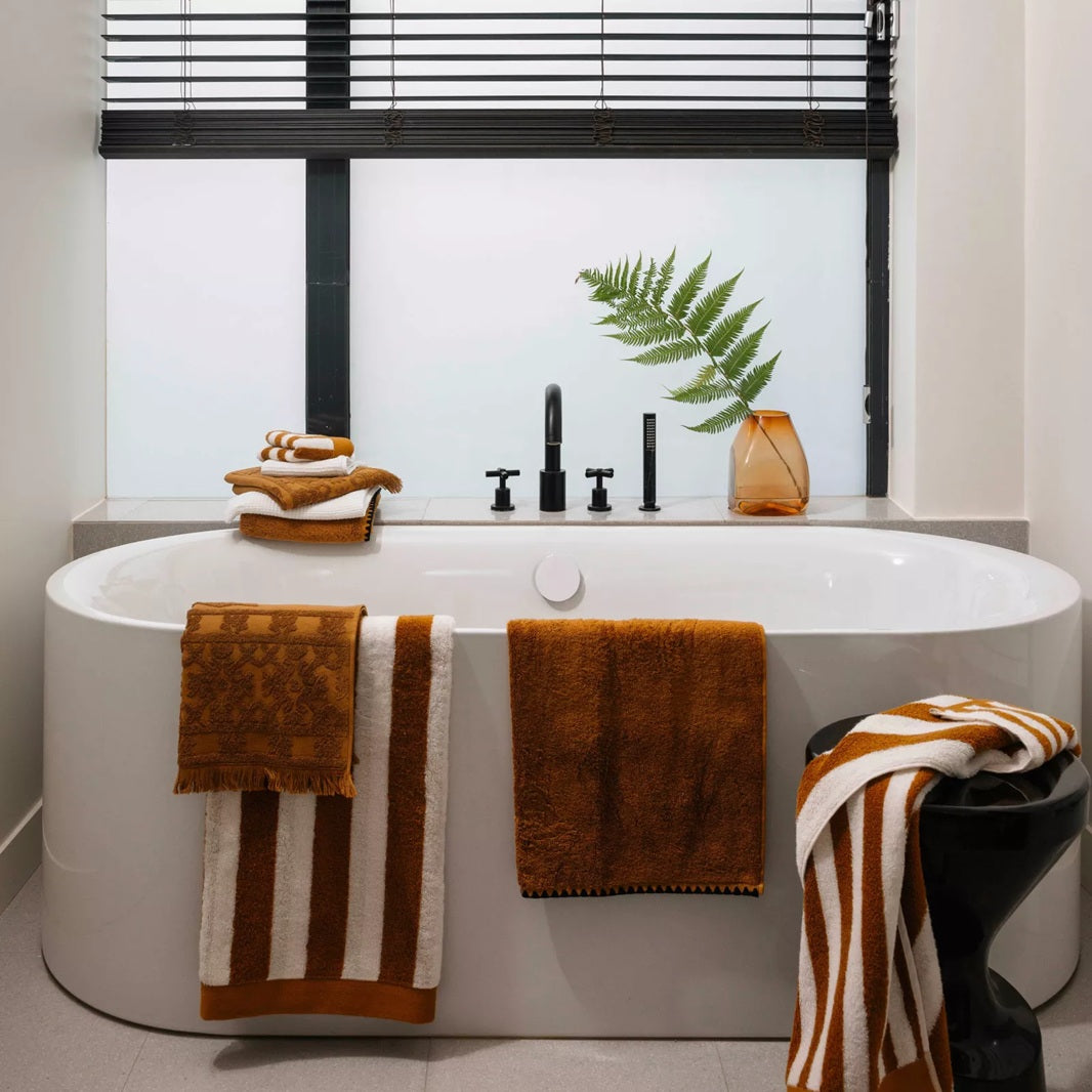 A modern freestanding bathtub in a bright bathroom setting, styled with brown and white towels, a black faucet, and a vase with a fern branch.