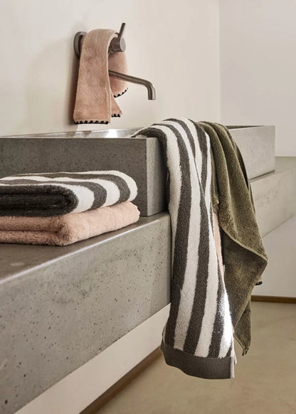 A set of folded and hanging towels in a minimalist bathroom setting with a concrete sink, including a striped white and dark gray towel, an olive green towel, and a folded pink towel.