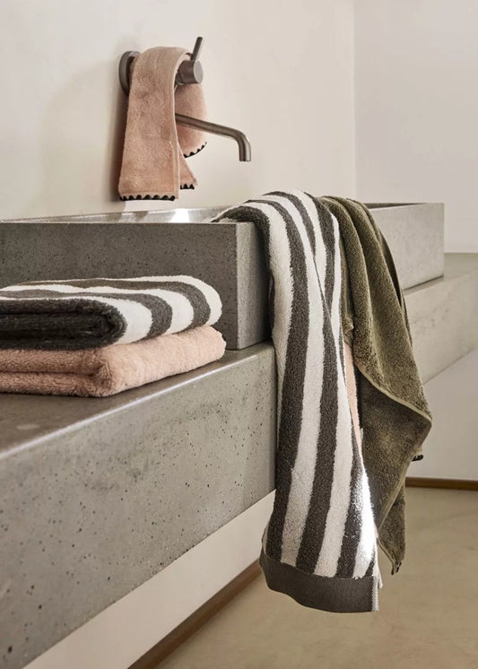 A set of folded and hanging towels in a minimalist bathroom setting with a concrete sink, including a striped white and dark gray towel, an olive green towel, and a folded pink towel.