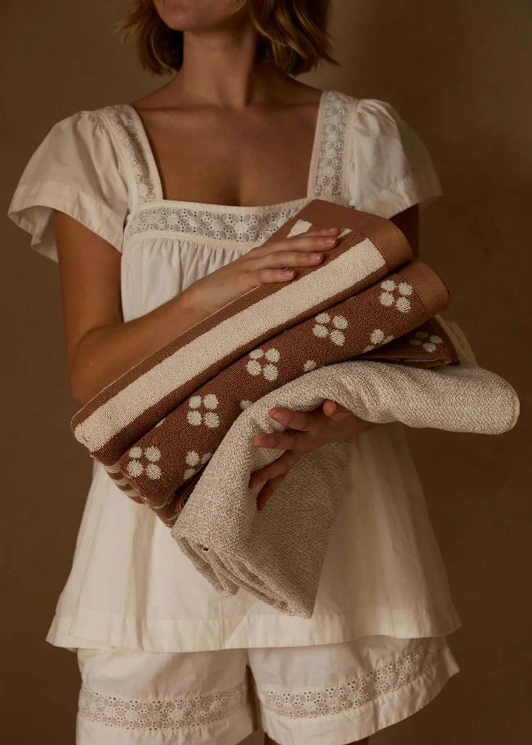 A woman in a cream-colored lace-trimmed top and shorts holds a folded set of textured towels in brown and cream with white floral motifs, set against a solid brown background.