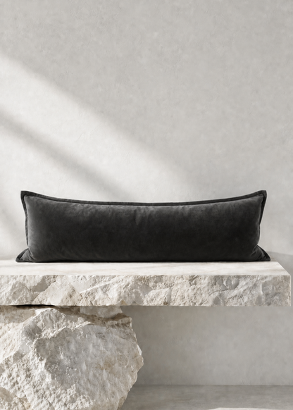 A long rectangular black velvet pillow displayed on a white stone bench against a textured light gray wall, with soft diagonal shadows cast across the scene.