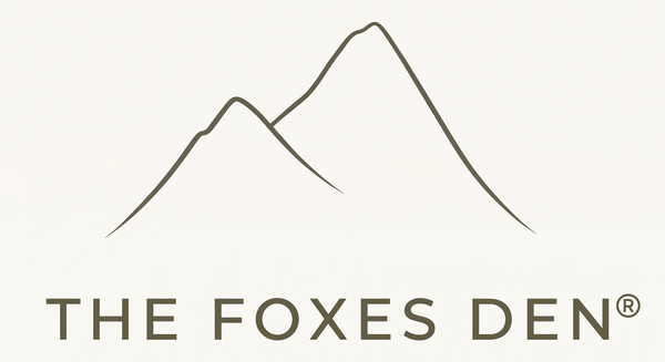 The Foxes Den logo featuring a minimalist black mountain line icon above the brand name in a dark brown serif font on a warm off-white background.