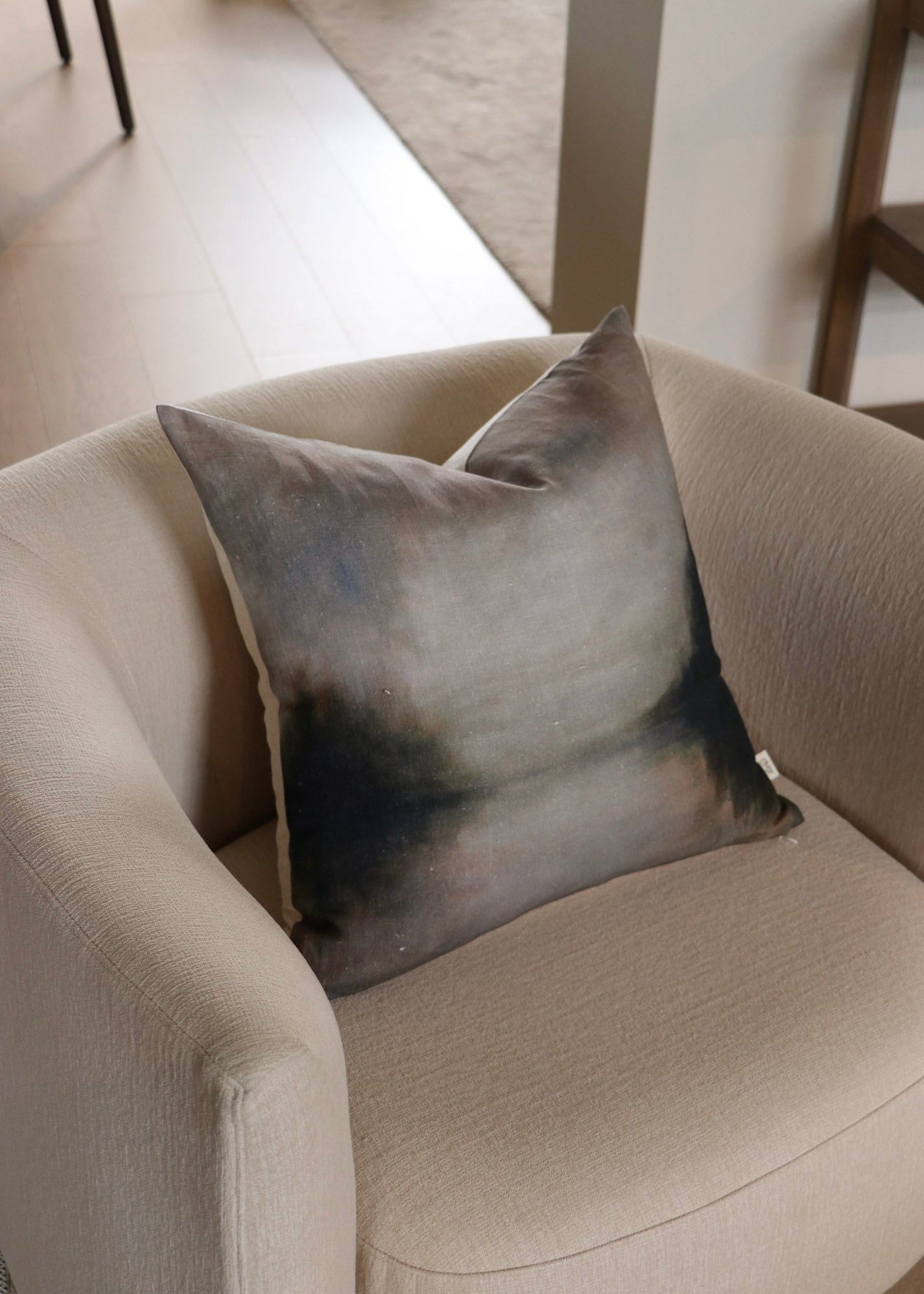 A square throw pillow with an abstract watercolor design in shades of gray, blue, and black, resting on a beige armchair in a modern living room setting.