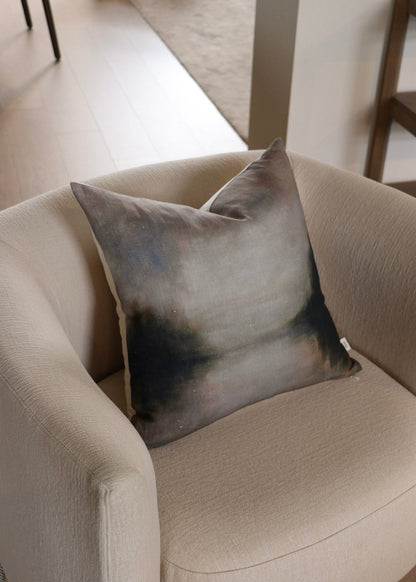 A square throw pillow with an abstract watercolor design in shades of gray, blue, and black, resting on a beige armchair in a modern living room setting.