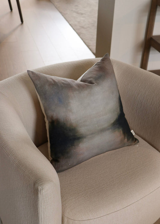 A square throw pillow with an abstract watercolor design in shades of gray, blue, and black, resting on a beige armchair in a modern living room setting.