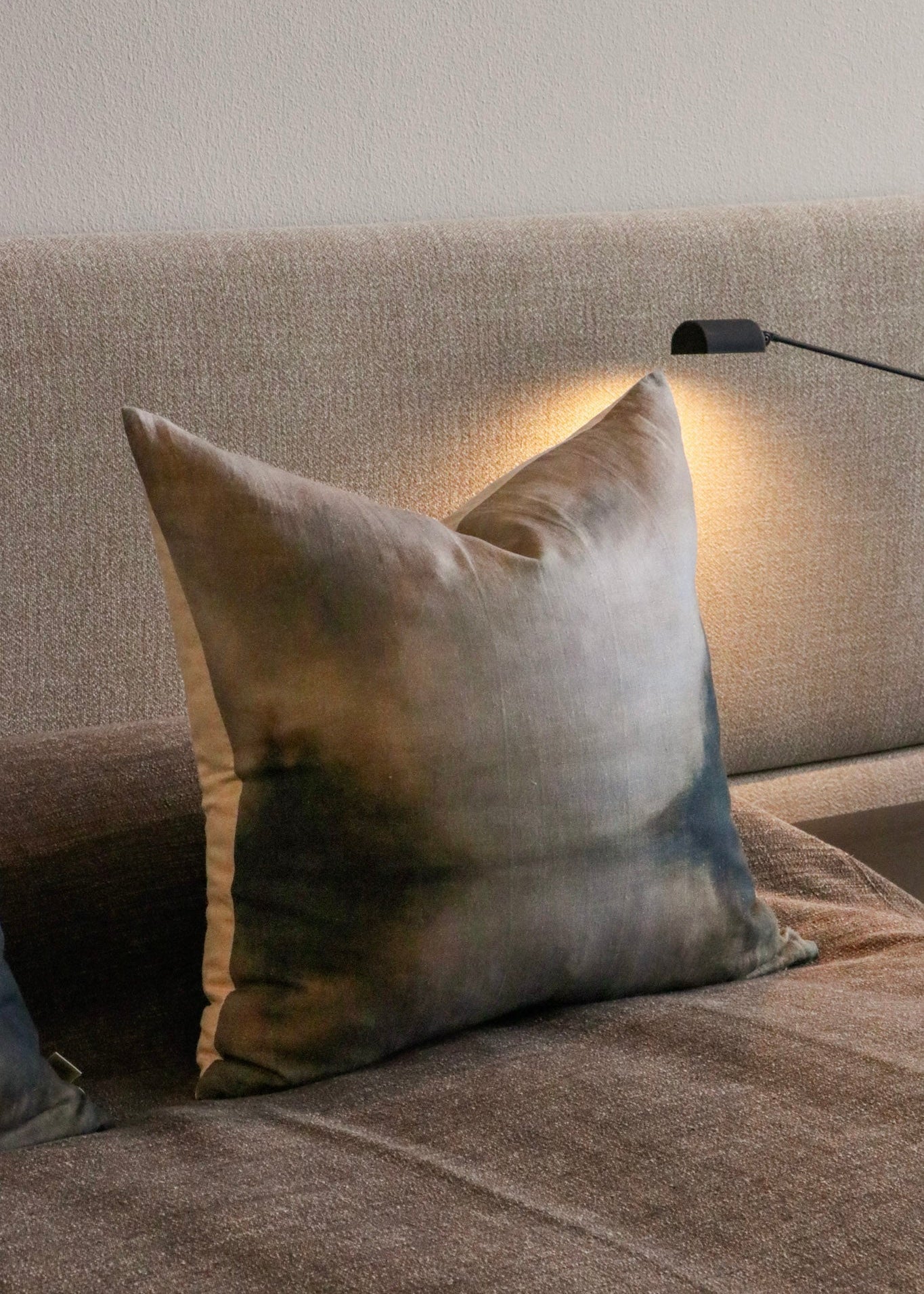 A square throw pillow with a textured linen-like finish, featuring a gradient color pattern of beige, gray, and dark charcoal, resting on a matching textured sofa under a warm light from a bedside lamp.