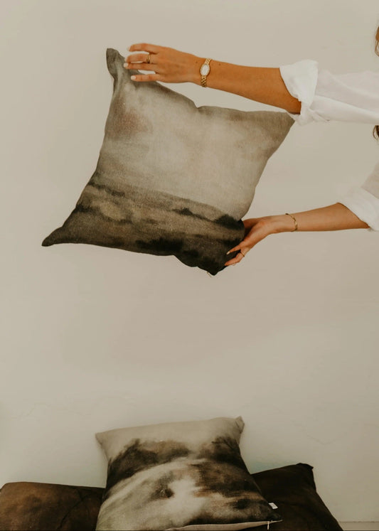 Person holding a decorative pillow with a neutral color palette against a plain background
