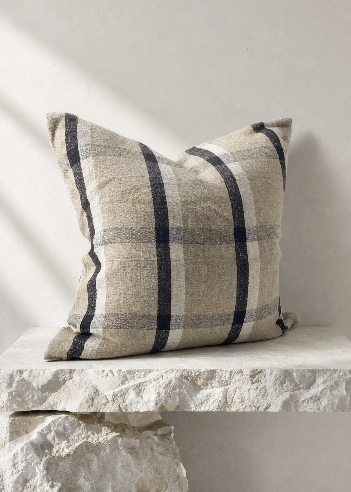 A square throw pillow with a beige and navy blue plaid pattern, resting on a rustic white stone bench against a neutral light gray wall.