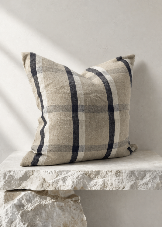 A square throw pillow with a beige and navy blue plaid pattern, resting on a rustic white stone bench against a neutral light gray wall.