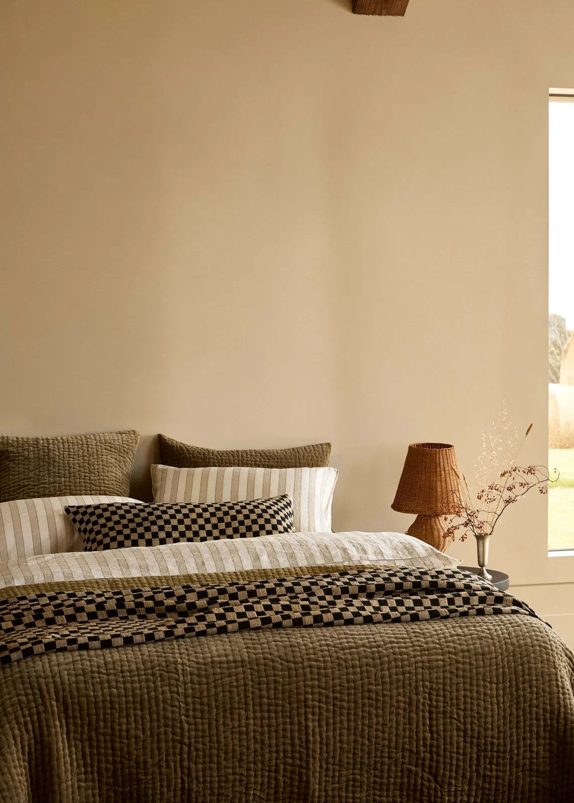 A styled bedroom scene featuring a bed dressed in textured olive green, cream, and black checkered bedding, set against a warm beige wall with a window and a lamp on a bedside table.