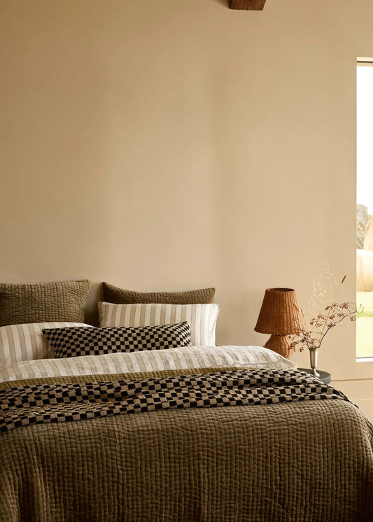A styled bedroom scene featuring a bed dressed in textured olive green, cream, and black checkered bedding, set against a warm beige wall with a window and a lamp on a bedside table.