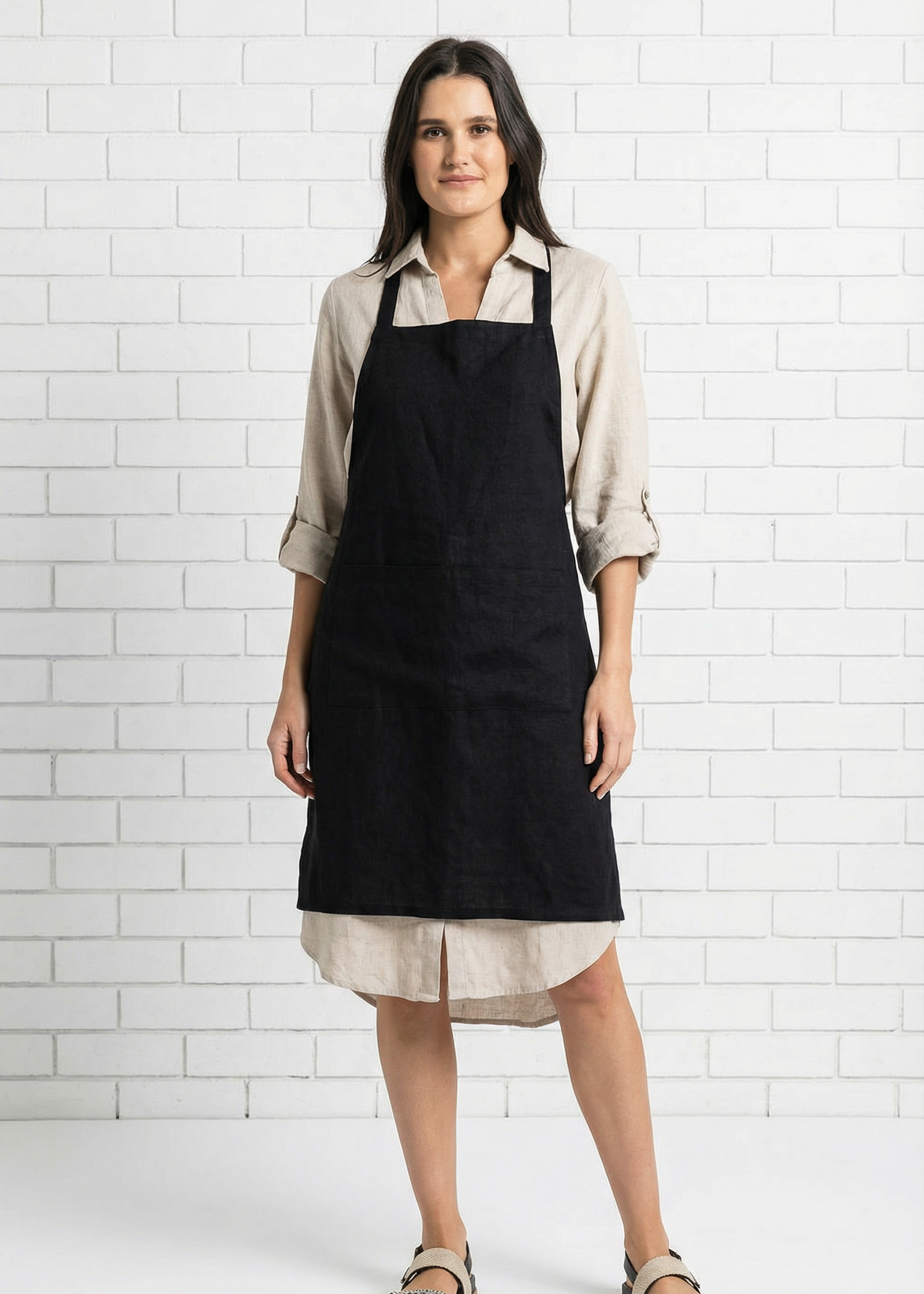 Woman wearing a black apron over a beige shirt and skirt against a white tiled wall.