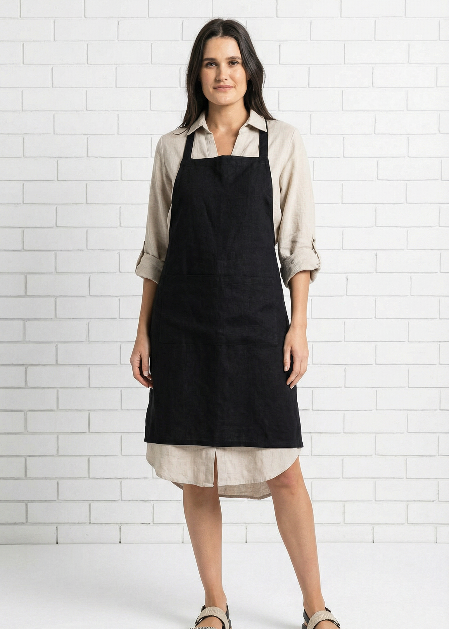 Woman wearing a black apron over a beige shirt and skirt against a white tiled wall.