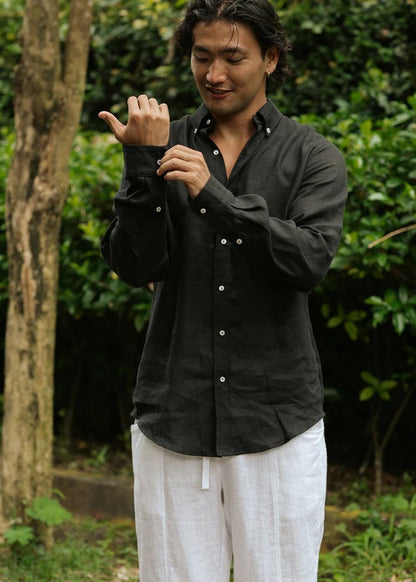Man wearing a black shirt and white pants standing outdoors with greenery in the background