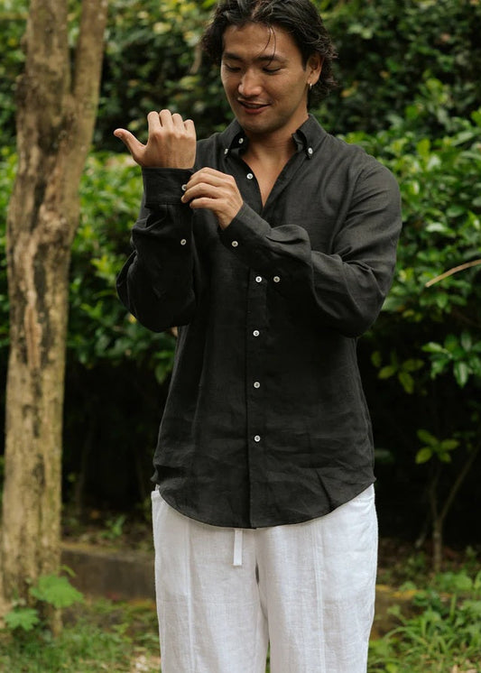 Man wearing a black shirt and white pants standing outdoors with greenery in the background
