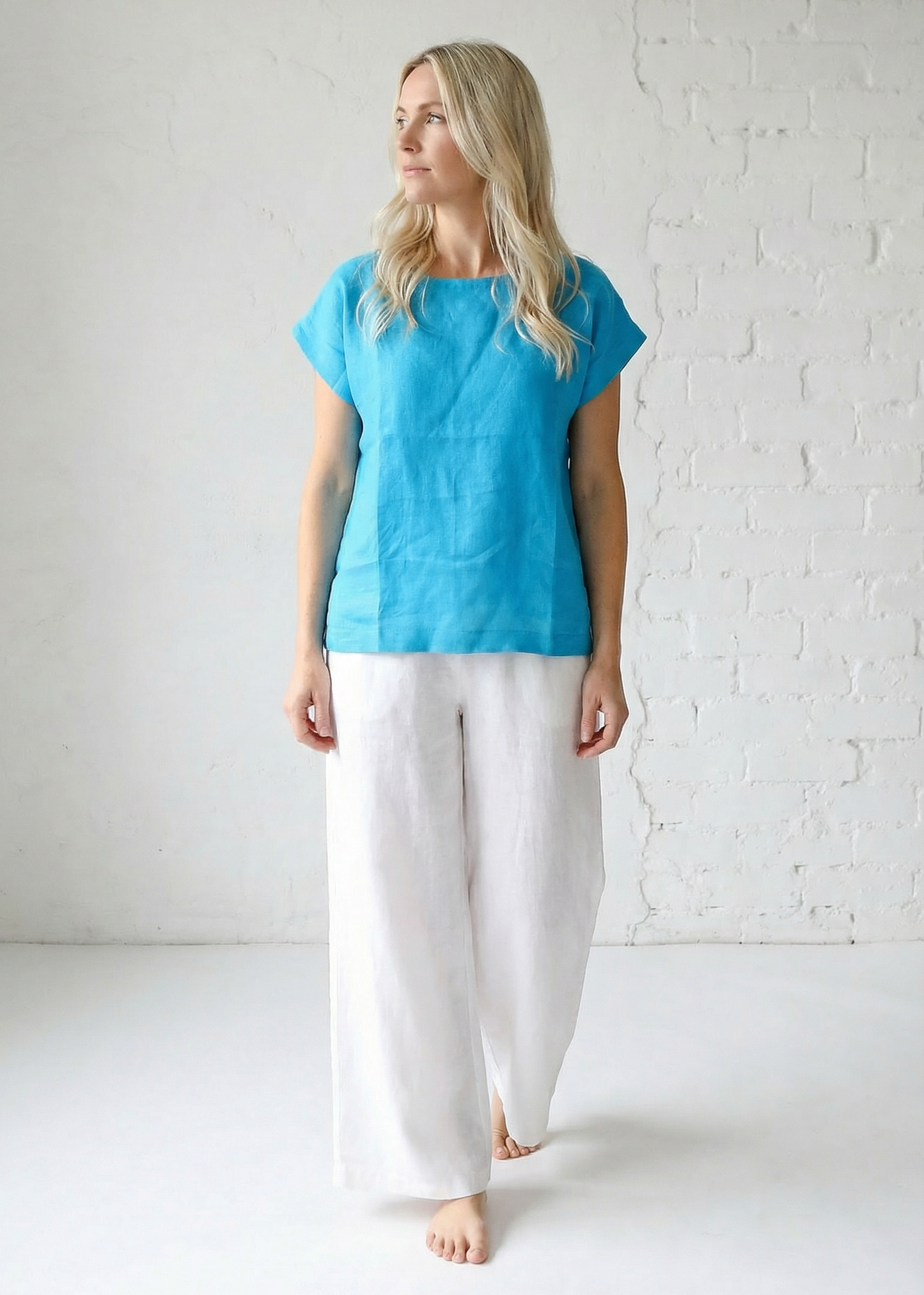 Woman wearing a blue top and white pants against a white brick wall.