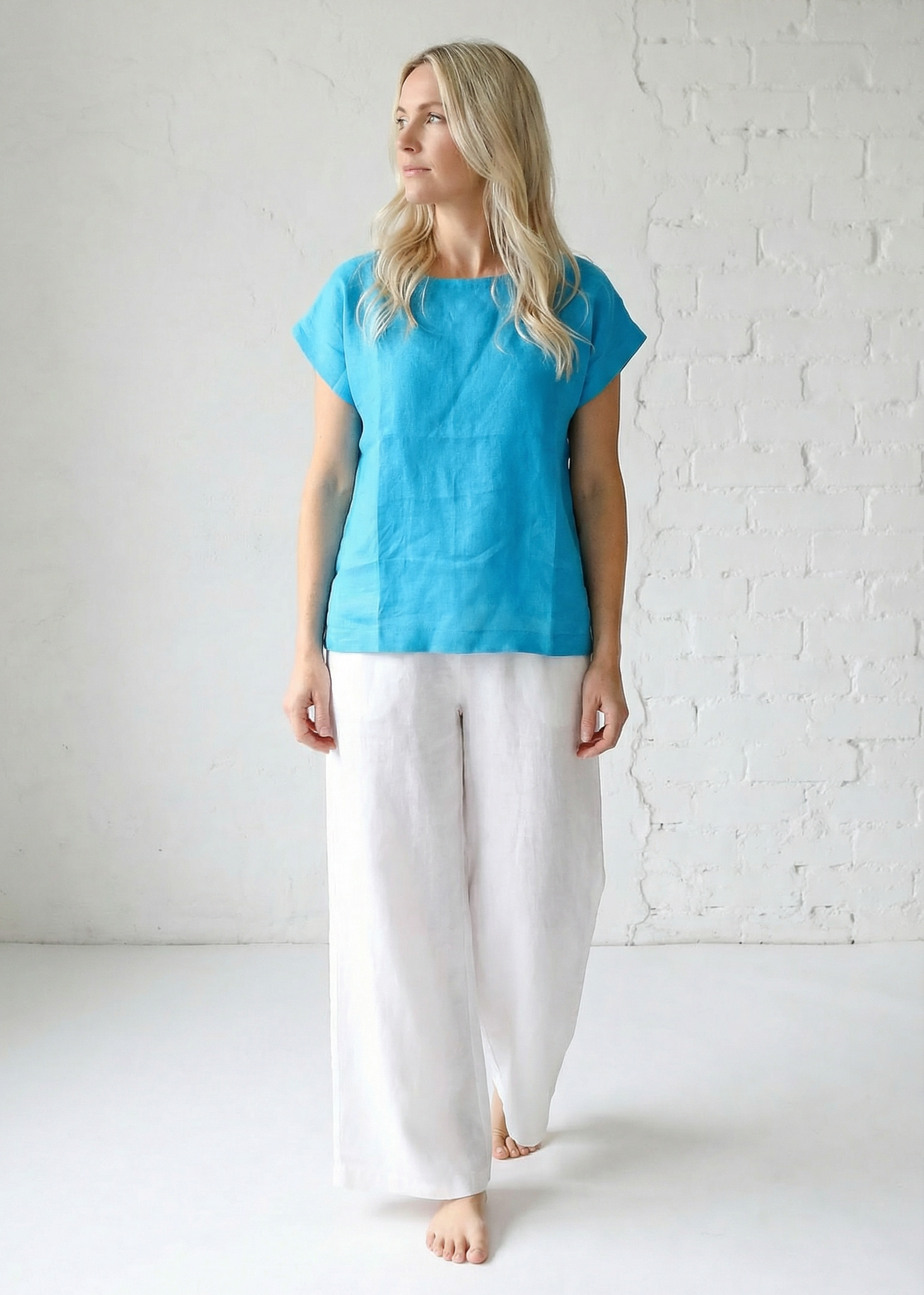Woman wearing a blue top and white pants against a white brick wall.