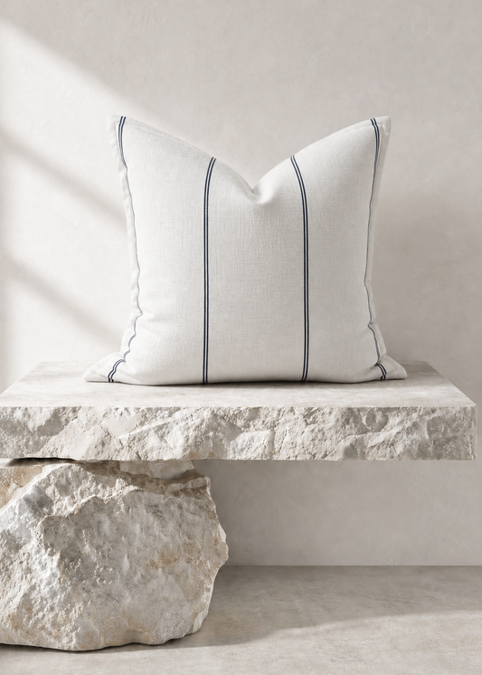 A square white throw pillow with two vertical navy blue stripes, styled on a rustic stone bench against a neutral beige wall.
