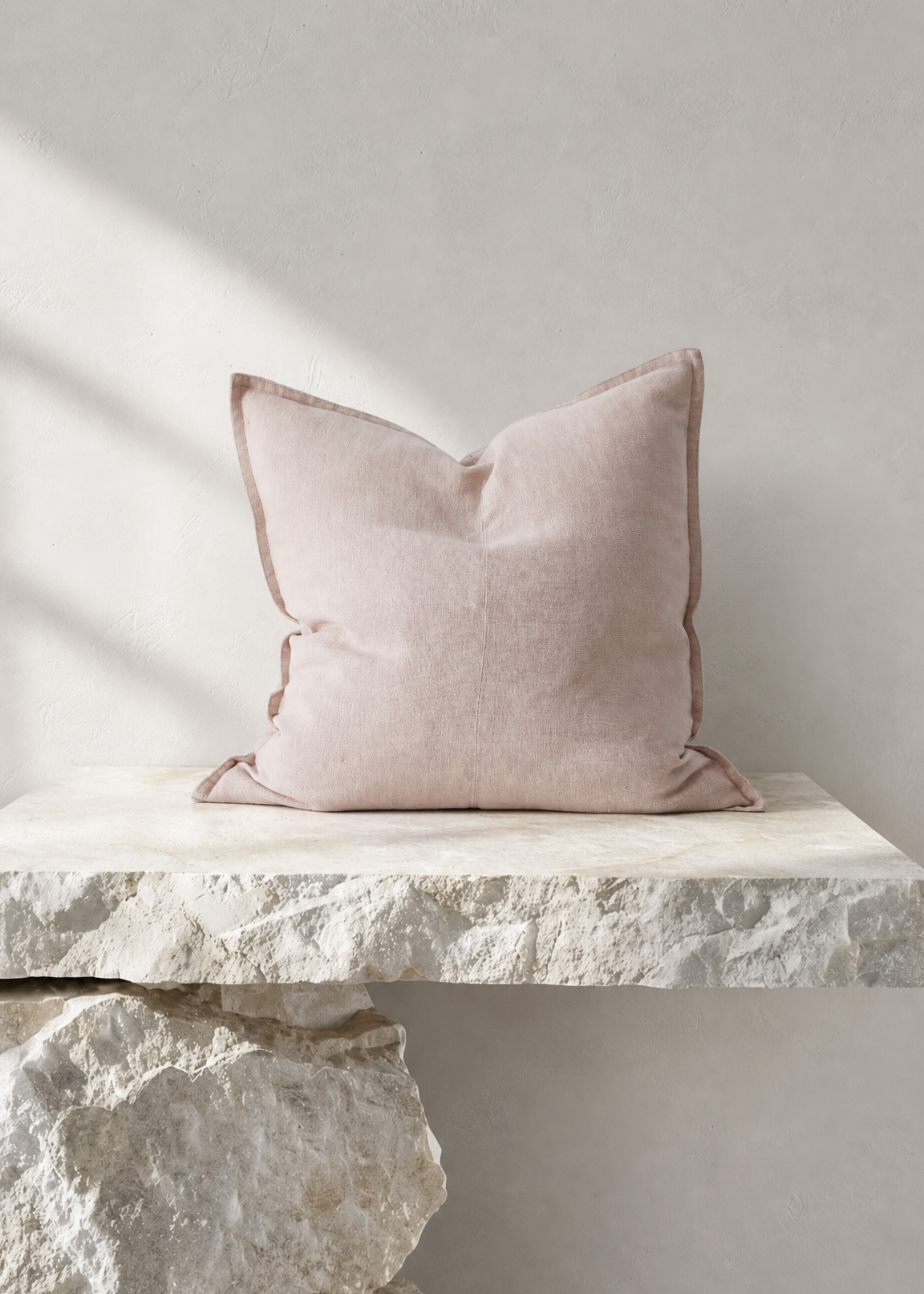 A square blush pink throw pillow with piped edges, resting on a rustic white stone ledge against a neutral light gray wall.