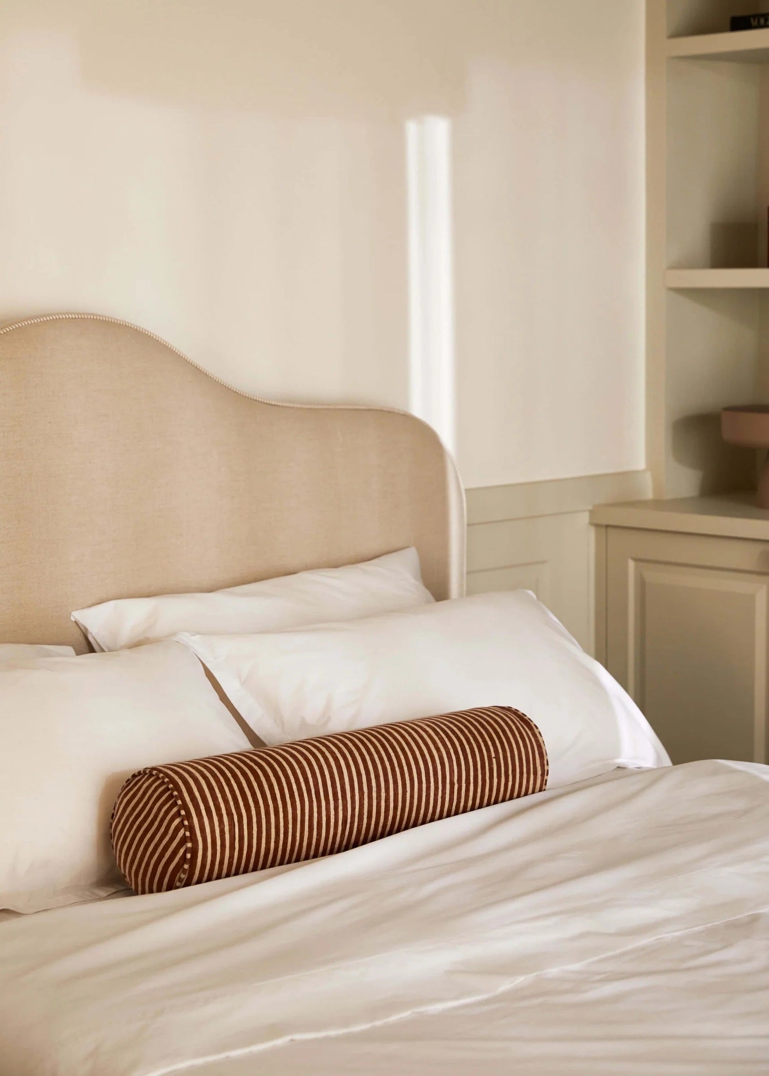 A hand block printed linen bolster cushion with a subtle stripe design, placed on a bed with a wooden headboard and shelves in the background.