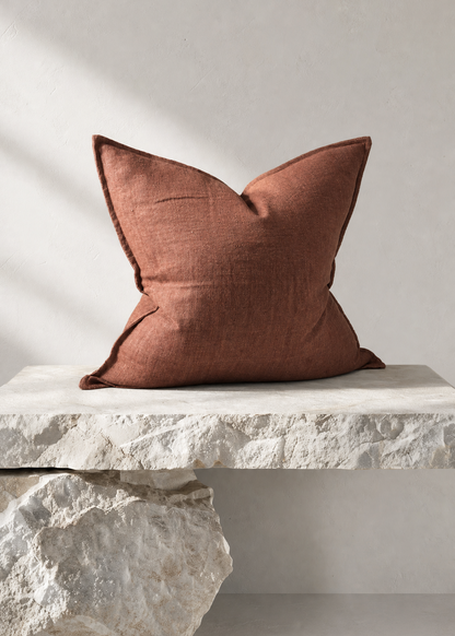 A square terracotta-colored linen throw pillow displayed on a rustic white stone bench against a neutral light gray wall.