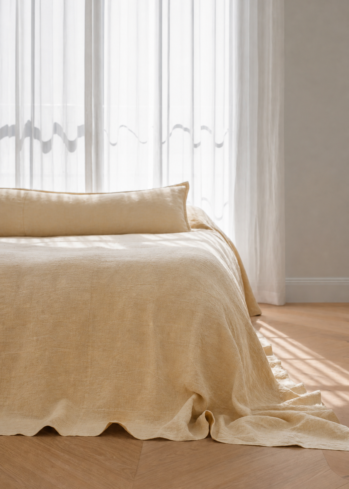 A beige linen throw blanket and matching pillow are draped over a bed in a softly lit bedroom with sheer white curtains and light wood flooring.